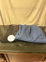 Folded blue Adidas sweatpants with logo on top, black fleece sweatpants beneath them.