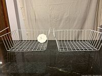 Two white wire baskets displayed side by side on black surface, showing overall structure and hooks.