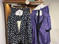 Two floral blouse and pleated skirt sets hanging side by side; one black with beige floral print, one purple with a smaller floral pattern.
