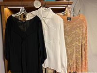 Three women’s dressy blouses hanging on plastic hangers on wooden rack. Left black blouse with V-neck, middle white blouse with collar and embroidery, right tan patterned semi-sheer blouse.