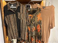 Three blouses and tunics hanging on a rack including black and brown geometric patterned blouse, brown and orange floral tunic, and plain brown blouse.