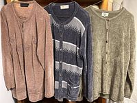 Image displaying three women’s long-sleeve cardigans hanging, one brown with floral embroidery, one grey with striped pattern and pockets, one green plush textured.
