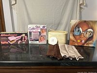 Vintage women's hairstyling tools lot on black surface with cream backdrop. Includes hairstyling devices, cosmetic bag, hosiery, and themed box.