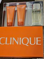 Open box showing the contents: two orange tubes of body wash, perfume spray bottle, and empty rollerball slot.