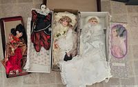 Five porcelain dolls displayed on a tiled floor. Left to right: boxed doll in pink dress, jester doll in red and black costume with white mask, Victorian-style doll in cream dress with bonnet, Victorian-style doll in white dress, geisha doll in red kimono.