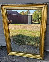 Front view of a rectangular mirror with a gold-tone ridged frame placed outdoors leaning against a light-colored wall reflecting grass and a shed.