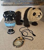 Photo showing panda figurine, Greek souvenir vase, and magnifier glass necklace together on a flat surface.