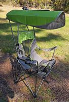 Green and gray portable folding camping chair set up outdoors with attached canopy overhead providing shade. Shows fabric details, cup holder, padded armrests, and metal frame structure.