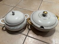 Two vintage soup tureens placed on floor tile, one with gold handles and the other with a green leaf pattern and two handles
