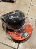 Six black snapback caps with orange brims and ventilation holes, stacked and encased in plastic packaging on a tiled floor.