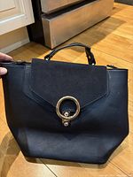 Front view of black backpack showing flap with suede-like panel and gold-tone circular clasp detail.