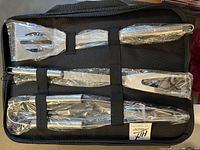 Three stainless steel BBQ utensils secured inside a black nylon carry case. Utensils include spatula, tongs and fork, all wrapped in protective plastic.