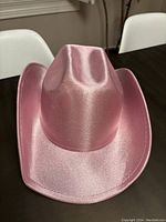 Front and slightly angled front view of shiny pink glittery cowgirl hat on dark table with white chairs in background.