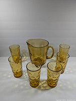 Front and angled view of one amber glass pitcher surrounded by six matching tall cups, all featuring a textured circle design on amber glass.