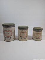 Front view of three nested metal cans of varying sizes decorated with a heart surrounded by floral accents and bows, with green lids featuring white polka dots.
