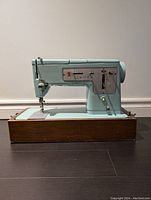 Front and side profile of vintage robin blue Singer sewing machine mounted on wooden carrying case base showing needle, presser foot and controls.