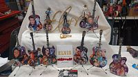 Full view of eight miniature Elvis Presley-themed guitars displayed on stands, with an Elvis gold-tone wall hanging behind them, the Elvis Aloha from Hawaii DVD set in front with some CDs