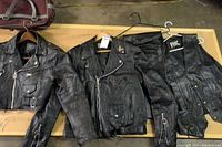 Two black leather jackets, black leather vest, and black leather chaps laid out