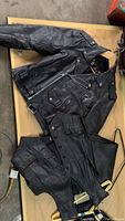 Full view of black leather motorcycle jacket, vest, and chaps laid out flat, showing front details, pockets, zippers, and snap buttons.