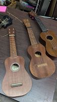 Top front view of three miniature wooden ukuleles showing bodies and necks, two without strings, one with some strings and a visible label 'GGA' inside sound hole.