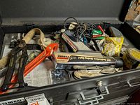 Photo of open top of black toolbox showing various hand tools including utility cutters, timing light, pliers, and other items.