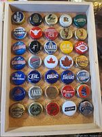 Top-down view of 30 assorted beverage bottle caps in a wooden and glass display box, arranged neatly on cork backing.