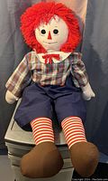 Full front view of a large Raggedy Andy cloth doll sitting upright showing its detailed outfit and red yarn hair.