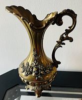 Photo of gold finish pitcher vase showing ornate handle, floral relief decoration, and scalloped rim.