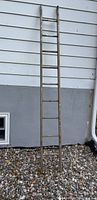 Photo of a tall, narrow wooden ladder towel holder leaning against a white and gray wall on a gravel surface. The ladder has eight rungs and a weathered, rustic finish.
