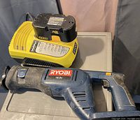 Blue and black Ryobi reciprocating saw with yellow 18V battery and charger laid on a gray surface.