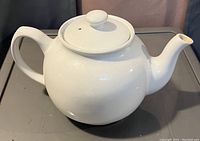 Photo of a white ceramic teapot with lid on a surface, showing side view with spout and handle visible.