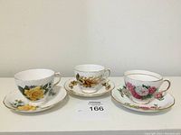 Three teacup and saucer sets shown from the front, displaying floral patterns with various colors and gold rims.