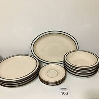 Photo showing the cream-colored Denby stoneware dishes with brown and gray banded edges, including stacks of dinner plates, saucers, bowls, and the platter in background.