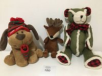 Photo of three holiday-themed plush stuffed animals: a tan dog with red and green Christmas hat and scarf, brown reindeer with antlers, and green bear with red plaid accents and scarf.
