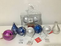 Nine assorted shatterproof Christmas ornaments in various colors and designs displayed on a white surface, with tags on some, including pink, blue, silver, and clear ornaments