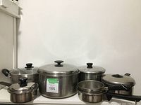 Six metal pots with lids, various sizes, arranged on a white surface. Largest pot lacks side handles; all have black knob lids.