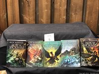 Five paperback Percy Jackson books by Rick Riordan lined up with front covers showing various mythical-themed illustrations such as mythical creatures and characters.