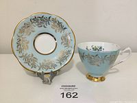 Teacup and scalloped saucer displayed side by side, showing gold gilt leaf pattern and blue background on both pieces.