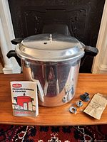 Reflective aluminum pressure canner with lid, black handles, instruction manual, and parts displayed on wooden surface.