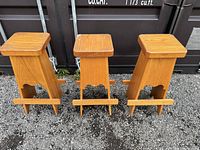 Photo showing three solid wood stools with square seats and footrests arranged outdoors on gravel in front of a dark storage container.