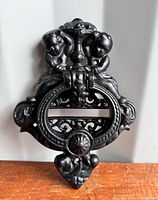 Front view of black antique cast iron door knocker with cherub figures and circular knocker ring.
