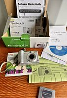 Canon PowerShot A75 camera shown with original packaging, user guides, software disc, and accessories on a wood surface.