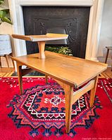 Angle view of the walnut wood mid-century side table showing the main top surface and upper raised tier, placed on a red patterned rug in front of a black fireplace.