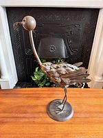 Front and side views of a tall metal bird sculpture with a curved neck and textured feathers, placed on a wooden surface in front of a fireplace.