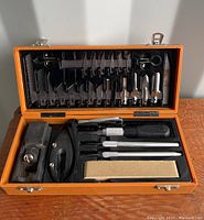 Opened orange metal case showing top compartment with multiple X-Acto blades and bottom compartment with handles, scissors, and sharpening stone.