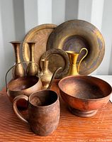 Photo showing the vintage assorted brass and copper items arranged on a wooden surface with two brass candlesticks, brass plates, several copper bowls, and brass pitchers.