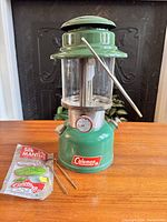 Green vintage Coleman Model 335 lantern shown with original packaging and silk mantles on wooden surface, fireplace in background.