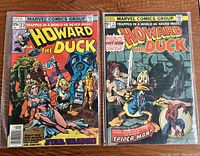 Both comic books laid flat showing cover art: Howard the Duck issue #23 with sci-fi themed cover and issue #1 with fantasy sword and Spider-Man theme.