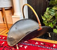 Side view showing the half-moon shape, black metal body with brass handle and feet on red carpet by fireplace.