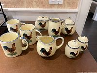 Photo showing the entire ceramic coffee set with rooster designs including mugs, creamers, sugar bowl with lid, and salt and pepper shakers arranged on a table.